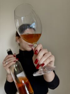 Natalia Zych – sommelierka Ta od Wina – degustuje wino pomarańczowe, trzymając kieliszek z bursztynowym winem i butelkę wina naturalnego